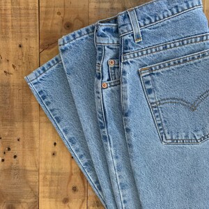 May include: Light blue denim jeans with classic five-pocket styling. The jeans are folded, showcasing the front pockets, belt loops, and a back pocket with a stitched design. The fabric has a faded appearance.