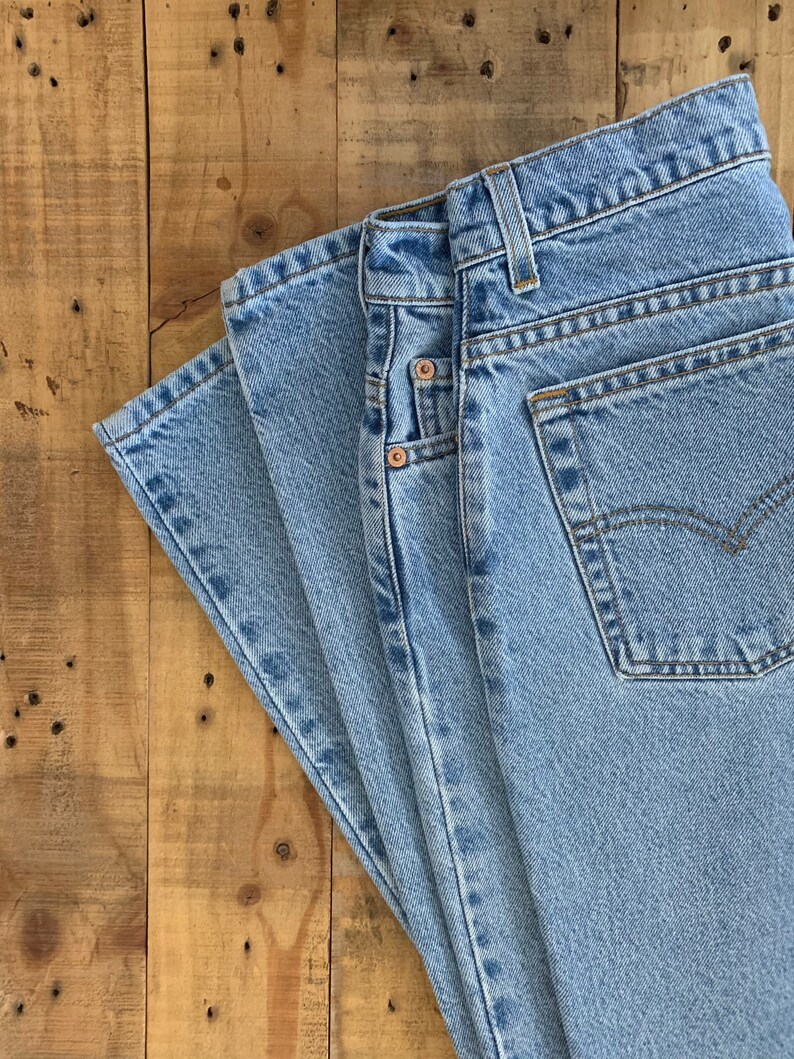 May include: Light blue denim jeans with classic five-pocket styling. The jeans are folded, showcasing the front pockets, belt loops, and a back pocket with a stitched design. The fabric has a faded appearance.