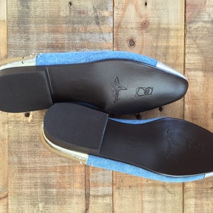 Puede incluir: Un par de zapatos de mezclilla azul con ribetes plateados y suelas de cuero marr&oacute;n. Las suelas est&aacute;n marcadas con la palabra "Amelia".