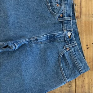 May include: Close-up of blue denim jeans with button and pocket details. The jeans are folded, showing the front pockets, belt loops, and a button closure. The denim has a classic, faded appearance.