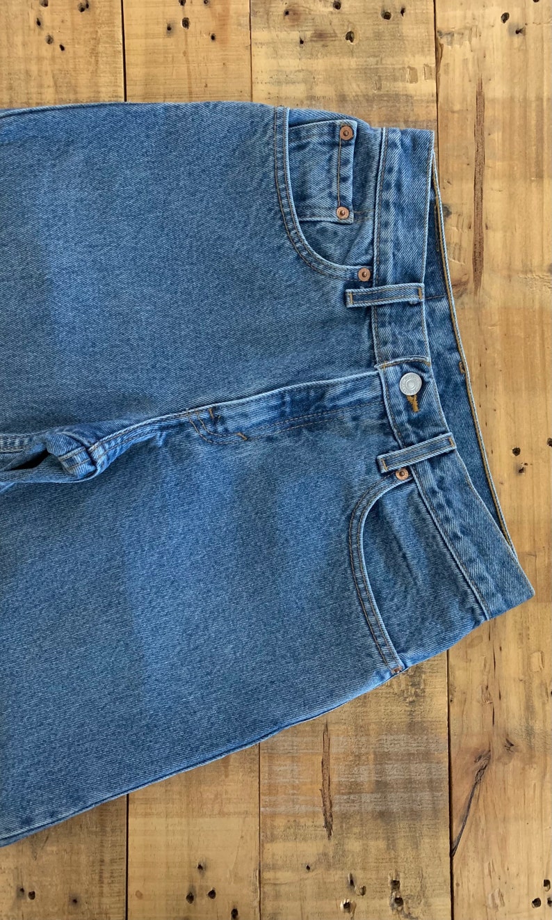 May include: Close-up of blue denim jeans with button and pocket details. The jeans are folded, showing the front pockets, belt loops, and a button closure. The denim has a classic, faded appearance.