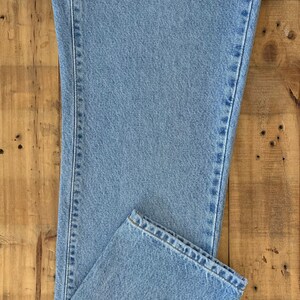May include: Light blue denim jeans, folded on a wooden surface. The jeans have a classic five-pocket design and a straight leg cut. The fabric appears to be a medium weight denim.