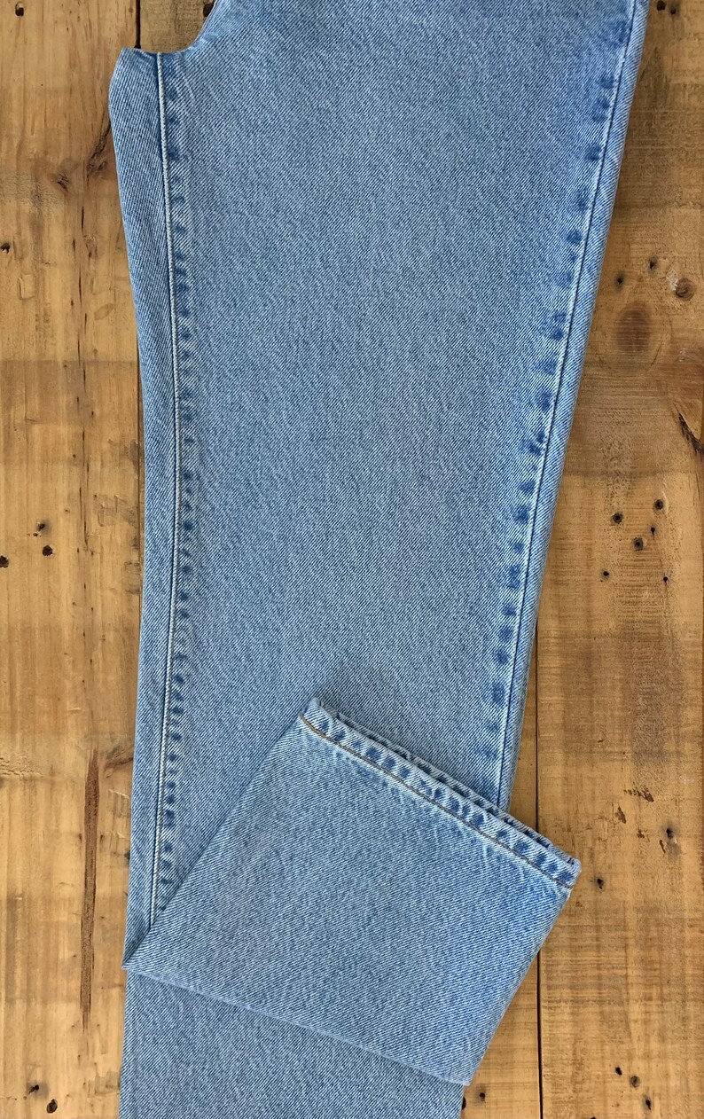 May include: Light blue denim jeans, folded on a wooden surface. The jeans have a classic five-pocket design and a straight leg cut. The fabric appears to be a medium weight denim.