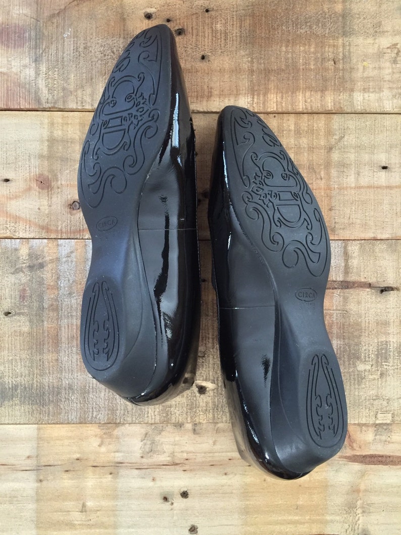 May include: A pair of black patent leather shoes with a decorative design on the soles. The shoes have a pointed toe and a low heel.