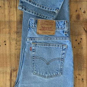 May include: Light blue Levi Strauss & Co. jeans with a classic five-pocket design. The jeans feature a leather patch with the Levi Strauss & Co. logo and the words "Personal Pair". The jeans are folded and laid flat on a wooden surface.