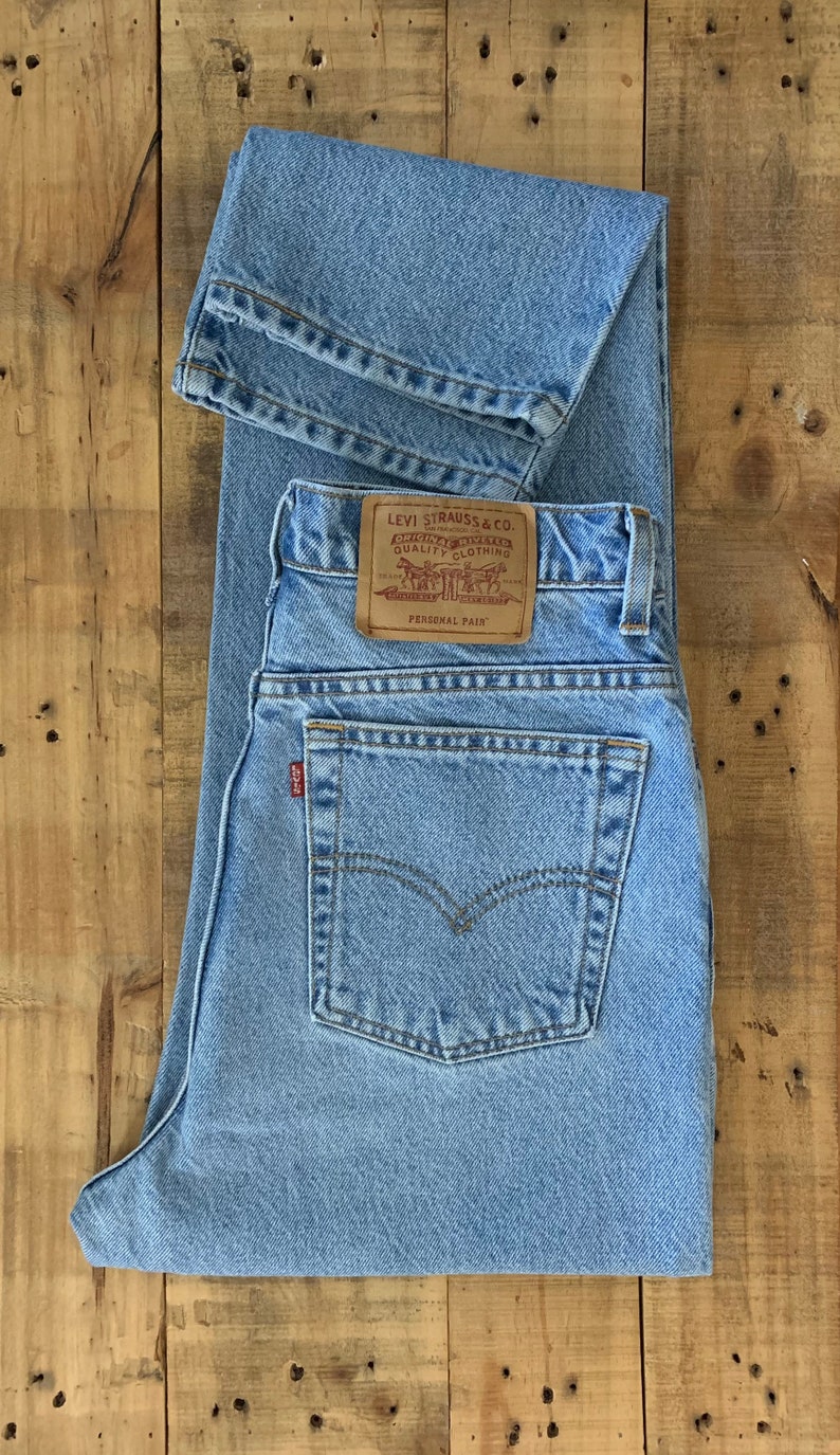 May include: Light blue Levi Strauss & Co. jeans with a classic five-pocket design. The jeans feature a leather patch with the Levi Strauss & Co. logo and the words "Personal Pair". The jeans are folded and laid flat on a wooden surface.