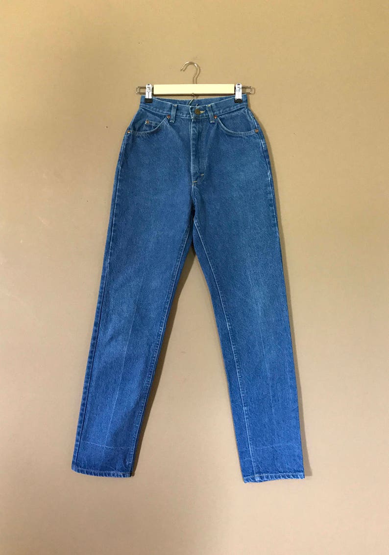 May include: A pair of blue denim jeans hanging on a wooden hanger against a brown background.