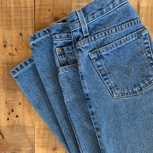 May include: Close-up of a pair of blue denim jeans, folded and stacked. The jeans have a classic five-pocket design, with a button and zip closure. The back pocket features a stitched design. The jeans are on a wooden surface.
