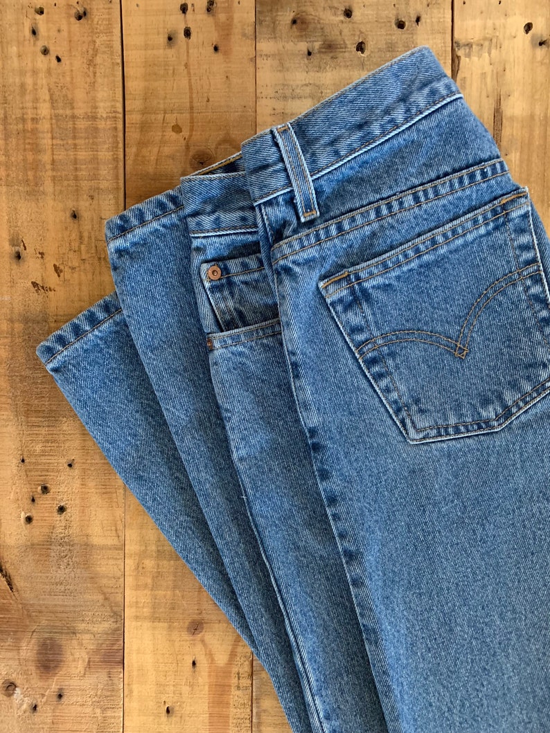 May include: Close-up of a pair of blue denim jeans, folded and stacked. The jeans have a classic five-pocket design, with a button and zip closure. The back pocket features a stitched design. The jeans are on a wooden surface.