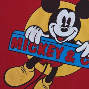 May include: Red t-shirt with a cartoon image of Mickey Mouse holding a blue sign that says "Mickey & Co."