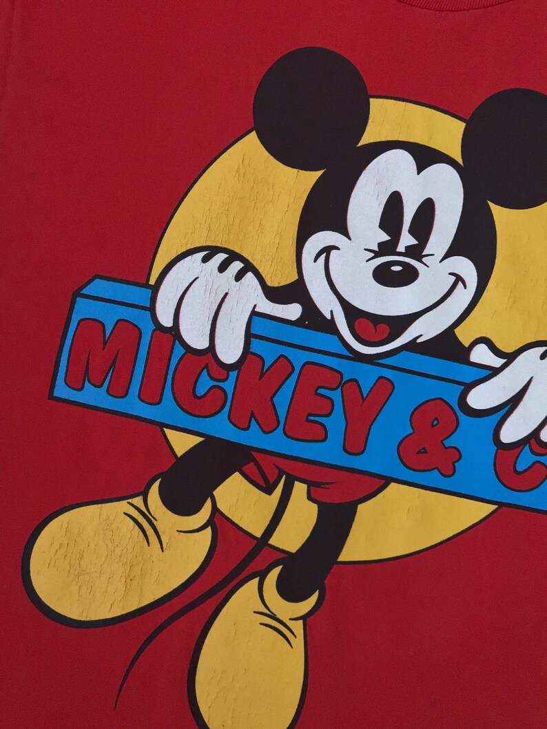 May include: Red t-shirt with a cartoon image of Mickey Mouse holding a blue sign that says "Mickey & Co."