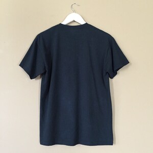 以下が含まれることがあります： ベージュの壁に白いハンガーにかけられた濃い青色のTシャツ