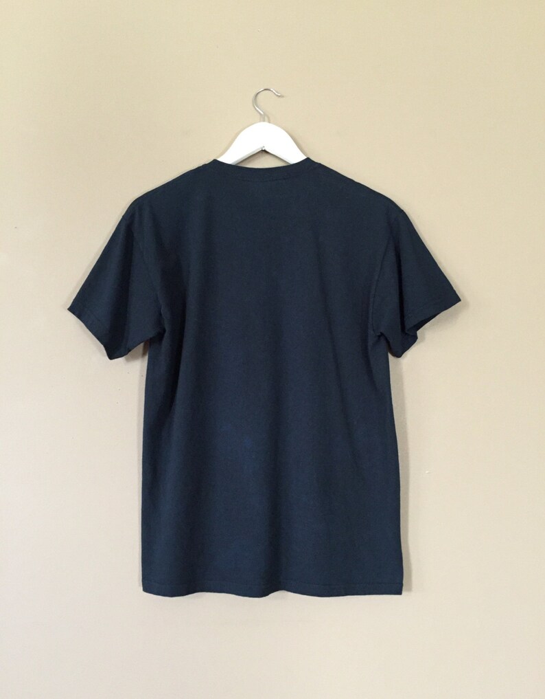 以下が含まれることがあります： ベージュの壁に白いハンガーにかけられた濃い青色のTシャツ