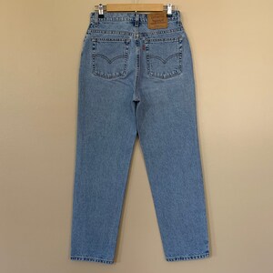 May include: Light blue denim jeans with a high waist, classic five-pocket design, and a leather patch on the back. The jeans are hanging on a wooden hanger. The back pockets have the signature Levi's stitching.