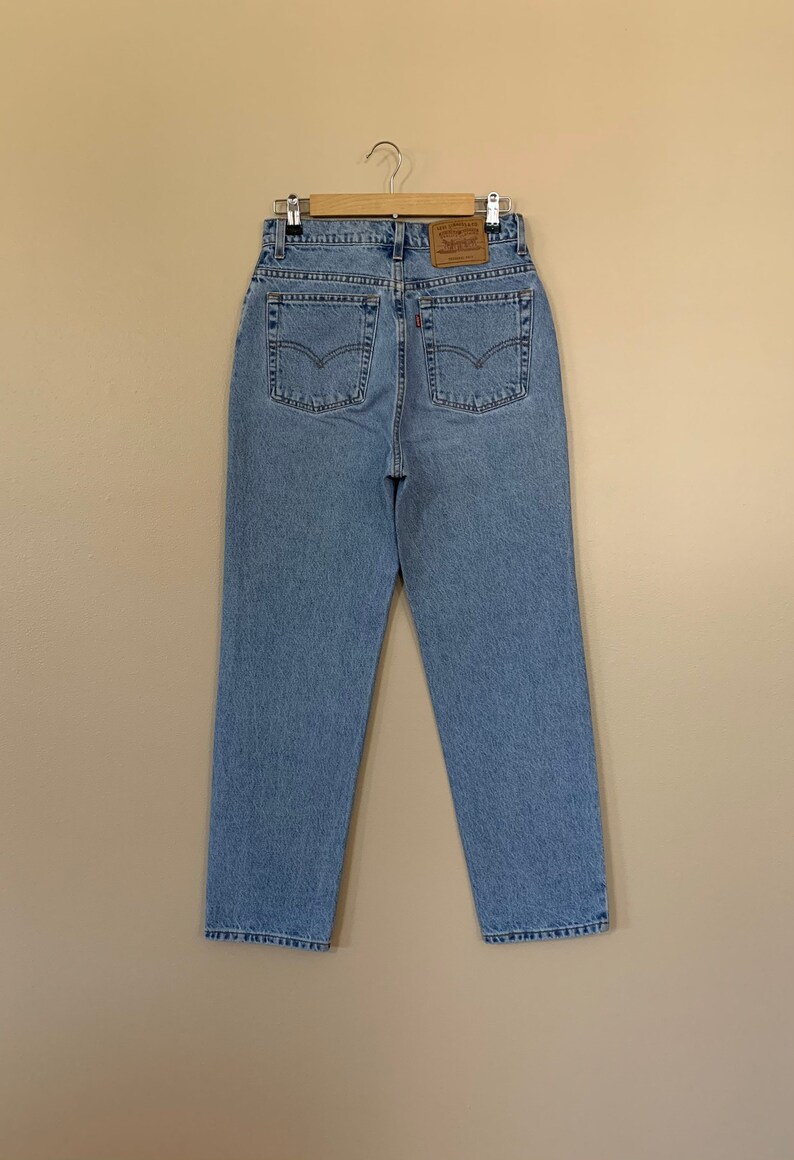 May include: Light blue denim jeans with a high waist, classic five-pocket design, and a leather patch on the back. The jeans are hanging on a wooden hanger. The back pockets have the signature Levi's stitching.