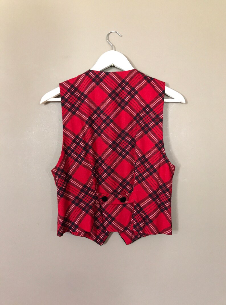 May include: A red and black plaid vest with a pointed collar and a button closure. The vest is sleeveless and has a slightly fitted silhouette.