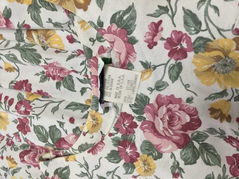 K&ouml;nnte beinhalten: Wei&szlig;er Stoff mit einem Blumendruck mit rosa und gelben Blumen. Der Stoff hat ein Etikett mit der Aufschrift "50% Polyester 50% Algodon Made in U.S.A. Hecho en E.U.A.  Cuta 12-109 Style 079622 Size Large/Grande"