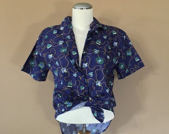 Puede incluir: Una camisa azul marino de manga corta con botones y un diseño estampado en blanco y verde. La camisa está anudada a la cintura.