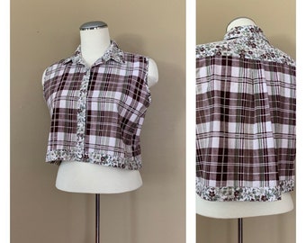 May include: A sleeveless cropped plaid shirt with a floral trim. The shirt is white, brown, and green plaid with a white button closure.
