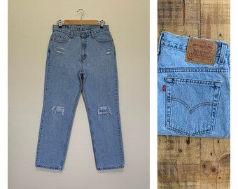 May include: Light blue denim jeans with distressing and rips. The jeans have a high waist and a straight leg fit. The back pocket has a red Levi's logo patch with the text "Levi Strauss & Co. Quality Clothing" and "501®" in white.