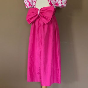 Puede incluir: Un vestido rosa con estampado floral blanco y un gran lazo en la espalda. El vestido tiene una abertura de ojo de cerradura en la espalda y mangas cortas abullonadas.
