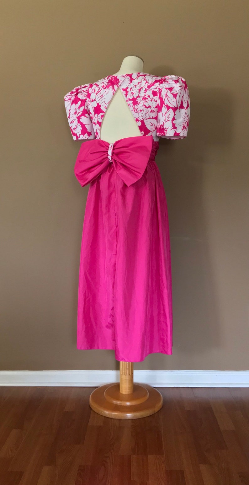 Puede incluir: Un vestido rosa con estampado floral blanco y un gran lazo en la espalda. El vestido tiene una abertura de ojo de cerradura en la espalda y mangas cortas abullonadas.