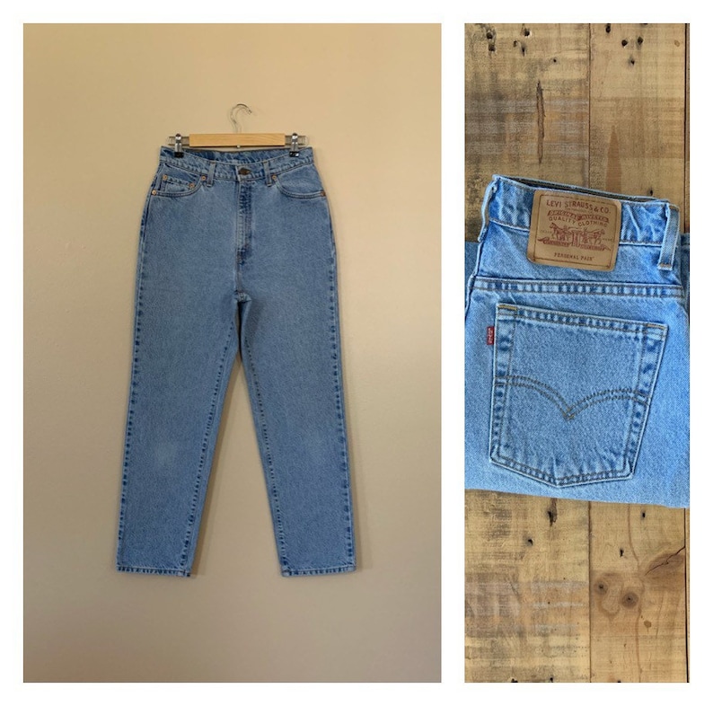 May include: Light blue denim jeans with a high waist, hanging on a wooden hanger. The jeans have a classic five-pocket design and a straight leg. A close-up shows the back pocket with a red tag and a leather patch.