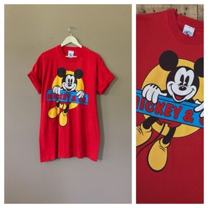 May include: Red t-shirt with a cartoon image of Mickey Mouse holding a blue and yellow sign that says "Mickey & Co."