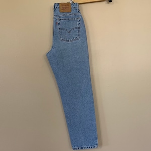 May include: Light blue denim jeans hanging on a wooden hanger. The jeans have a classic five-pocket design and a brown leather patch on the back. The jeans are a light wash and appear to be vintage.