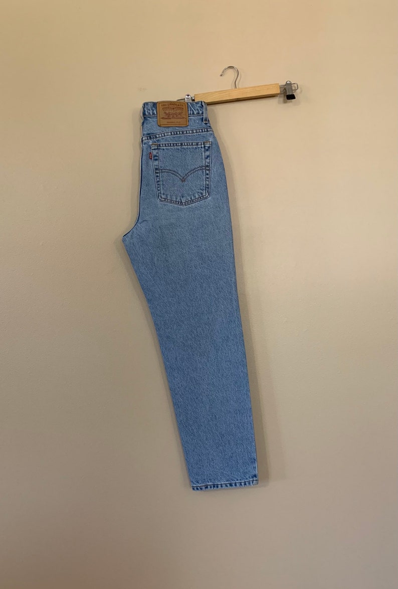 May include: Light blue denim jeans hanging on a wooden hanger. The jeans have a classic five-pocket design and a brown leather patch on the back. The jeans are a light wash and appear to be vintage.