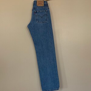 May include: Light blue denim jeans hanging on a wooden hanger. The jeans have a classic five-pocket design, a red Levi's tag, and a leather patch. The jeans are a straight-leg style.
