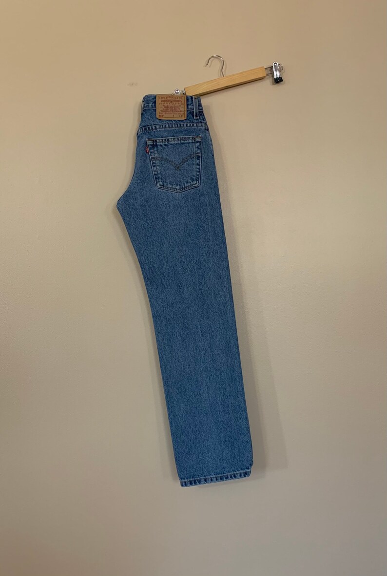 May include: Light blue denim jeans hanging on a wooden hanger. The jeans have a classic five-pocket design, a red Levi's tag, and a leather patch. The jeans are a straight-leg style.