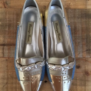 Puede incluir: Un par de zapatos de mujer plateados y dorados con una parte superior de denim azul. Los zapatos tienen una punta puntiaguda y una cadena plateada decorativa en la parte delantera. Los zapatos est&aacute;n etiquetados como "Carriage Court."