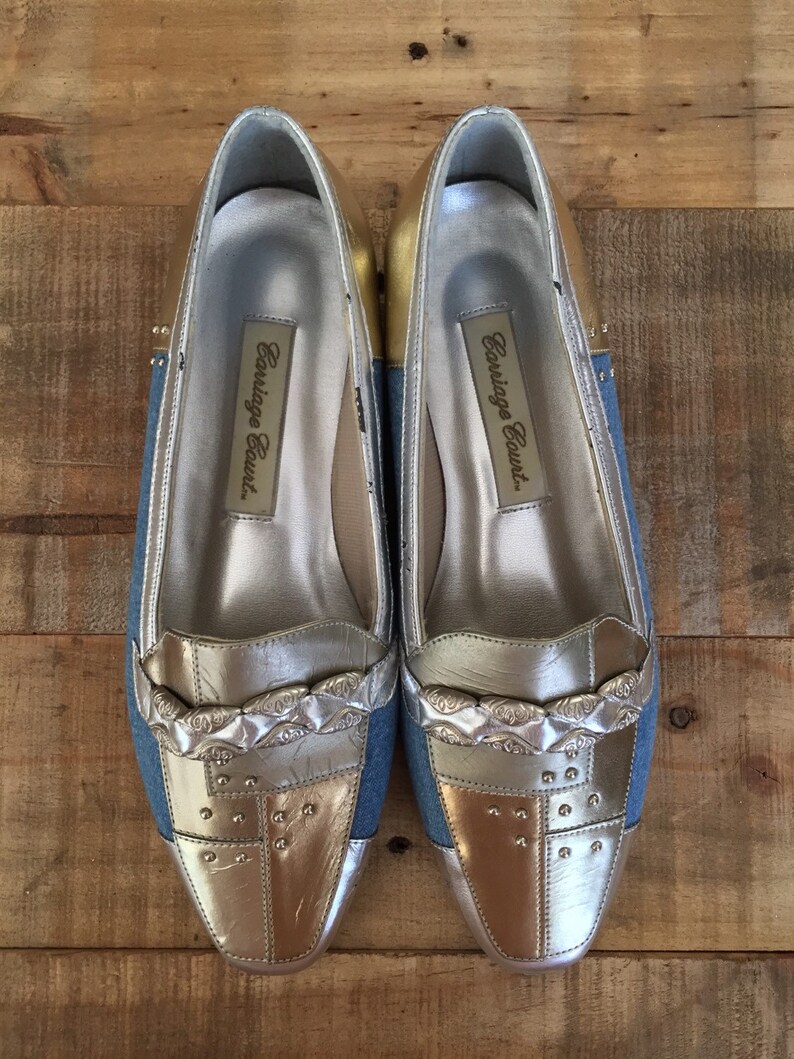 Puede incluir: Un par de zapatos de mujer plateados y dorados con una parte superior de denim azul. Los zapatos tienen una punta puntiaguda y una cadena plateada decorativa en la parte delantera. Los zapatos est&aacute;n etiquetados como "Carriage Court."