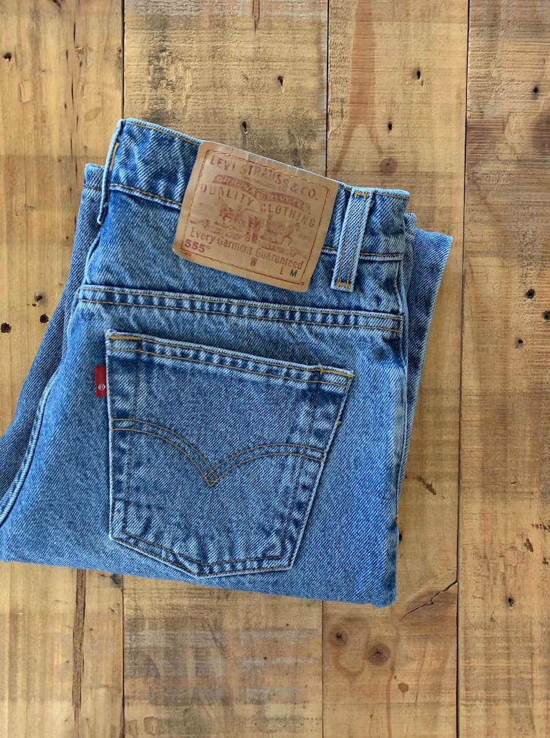May include: A pair of blue denim jeans with a brown leather Levi Strauss & Co. patch on the back pocket. The patch has the text "Original Quality Clothing" and "Every Garment Guaranteed" with the size 555 and W LM.