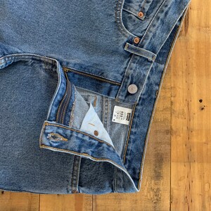 May include: Close-up of light blue denim jeans. The jeans have a button closure, a zip, and classic five-pocket styling. The Levi's label is visible. The jeans are on a wooden surface.