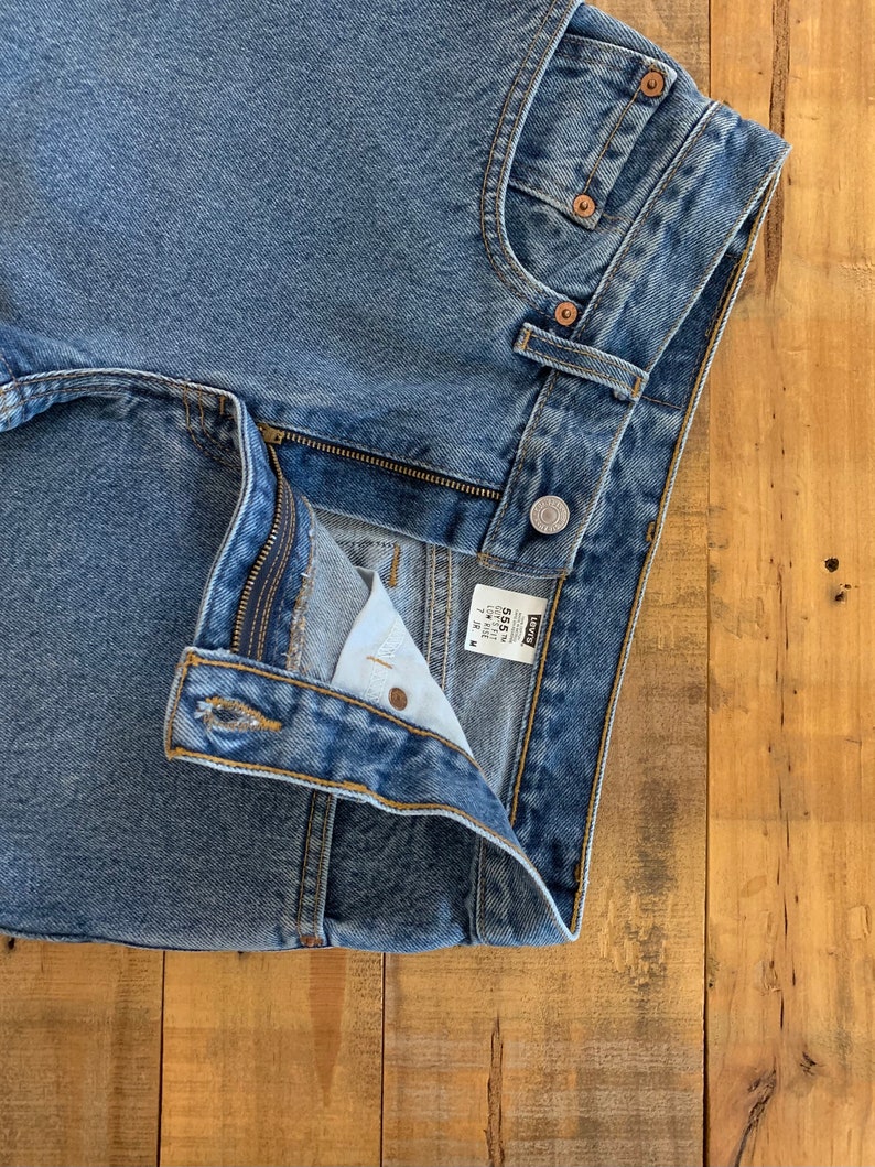 May include: Close-up of light blue denim jeans. The jeans have a button closure, a zip, and classic five-pocket styling. The Levi's label is visible. The jeans are on a wooden surface.