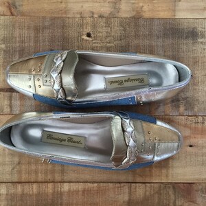 Puede incluir: Un par de mocasines plateados y dorados con un acento de mezclilla azul. Los mocasines tienen un detalle de correa trenzada y est&aacute;n etiquetados como "Carriage Court."