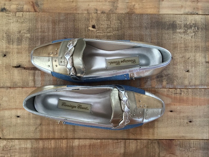 Puede incluir: Un par de mocasines plateados y dorados con un acento de mezclilla azul. Los mocasines tienen un detalle de correa trenzada y est&aacute;n etiquetados como "Carriage Court."