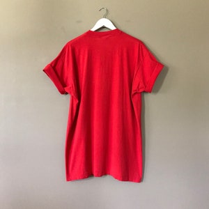 May include: A red short-sleeved t-shirt hanging on a white hanger against a beige wall.