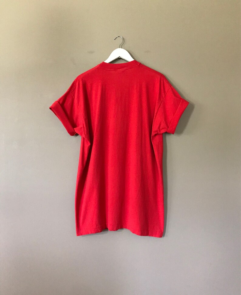 May include: A red short-sleeved t-shirt hanging on a white hanger against a beige wall.