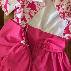 Puede incluir: Un vestido rosa con estampado floral blanco y un gran lazo rosa en la espalda. El vestido tiene un forro blanco y una cremallera en la espalda.