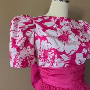 Puede incluir: Un vestido de estampado floral rosa y blanco con un gran lazo en la cintura. El vestido tiene mangas abullonadas y un corpi&ntilde;o ajustado.