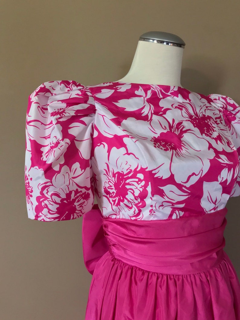 Puede incluir: Un vestido de estampado floral rosa y blanco con un gran lazo en la cintura. El vestido tiene mangas abullonadas y un corpi&ntilde;o ajustado.