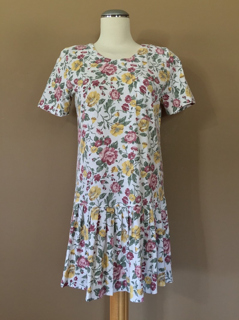 K&ouml;nnte beinhalten: Ein wei&szlig;es Kleid mit einem Blumendruck mit roten, gelben und gr&uuml;nen Blumen. Das Kleid hat kurze &Auml;rmel und einen gerafften Rock.