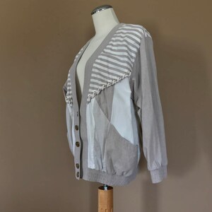 May include: A beige cardigan with a striped pattern on the upper back and a patchwork design on the front. The cardigan has a button closure and long sleeves.