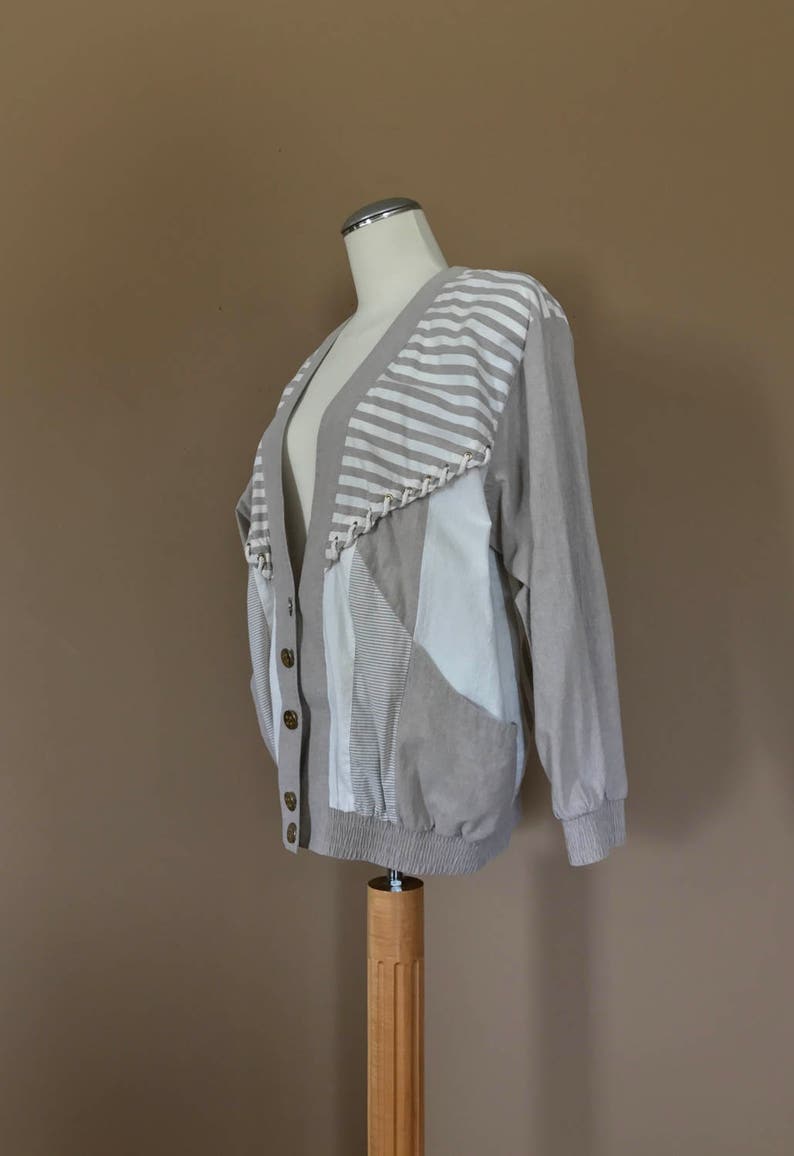 May include: A beige cardigan with a striped pattern on the upper back and a patchwork design on the front. The cardigan has a button closure and long sleeves.