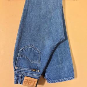 May include: A pair of blue denim jeans with a brown leather Lee label. The jeans are folded and hanging on a wooden hanger.