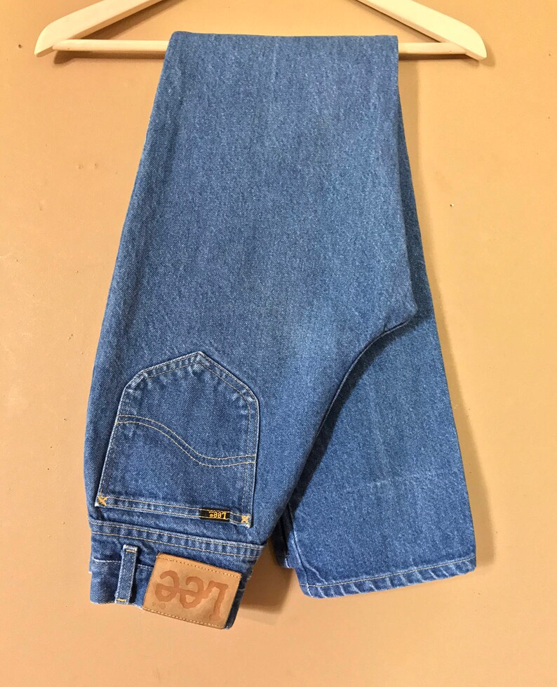 May include: A pair of blue denim jeans with a brown leather Lee label. The jeans are folded and hanging on a wooden hanger.