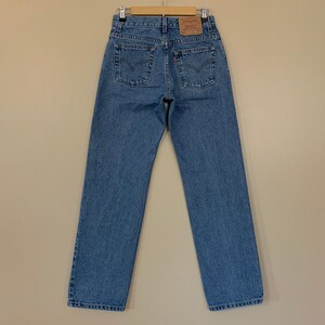 May include: Light blue denim jeans hanging on a wooden hanger. The jeans have a classic five-pocket design, a straight leg, and a brown leather patch on the back. The Levi's brand label is visible. The jeans are a medium wash.